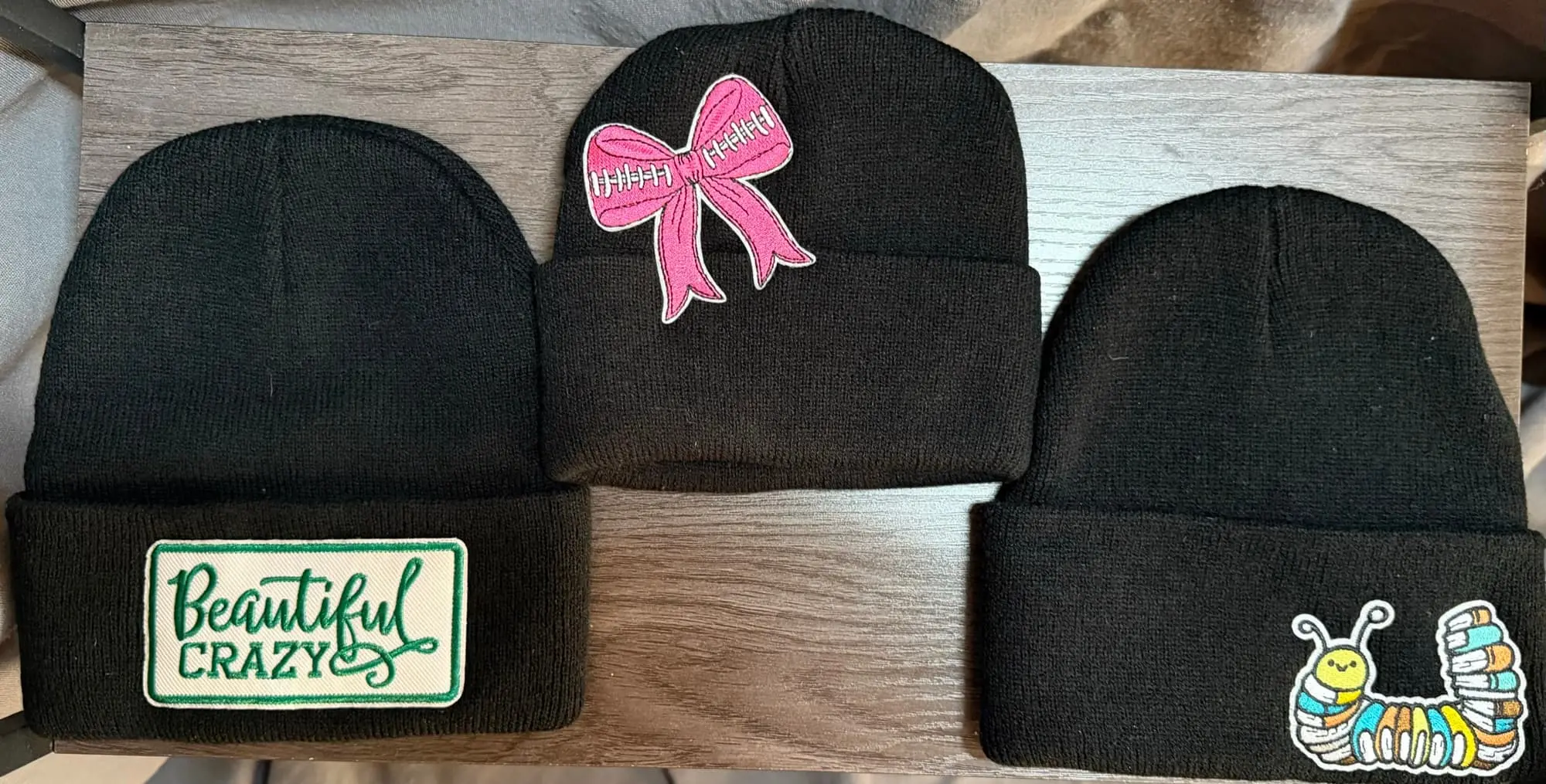Three black knit beanies laid flat: Beautiful Crazy, a pink football bow and a rainbow caterpillar