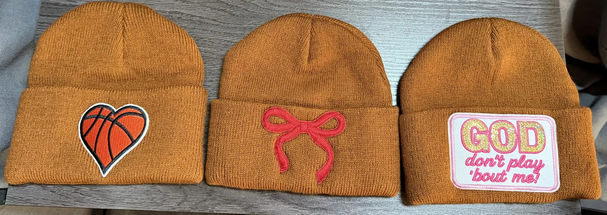 Three brown knit beanies: a basketball heart, a red bow and a GOD Don’t Play ’Bout Me patch