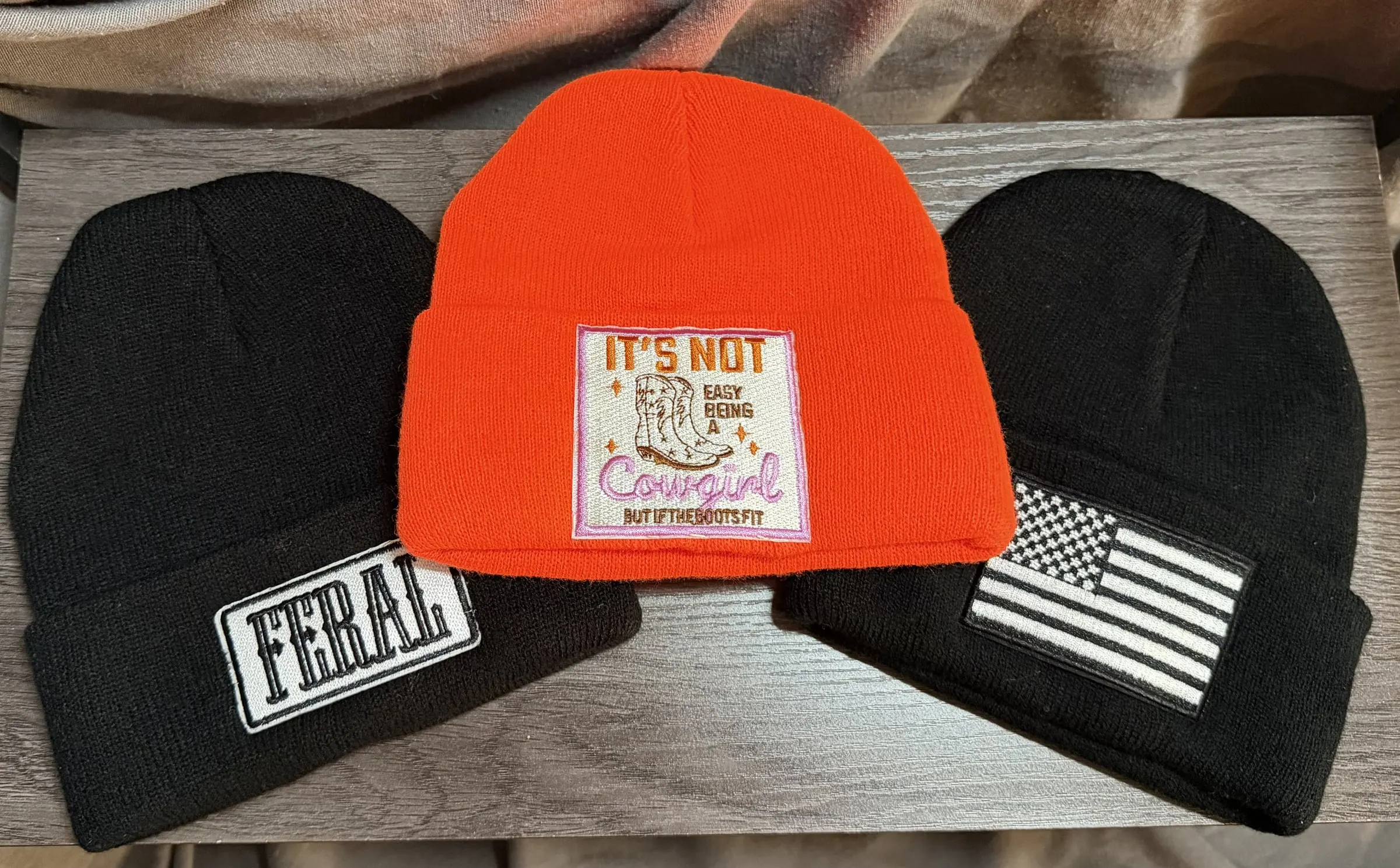 Three knit beanies: a black Feral patch, an orange It’s Not Easy Being a Cowgirl patch and a black flag patch