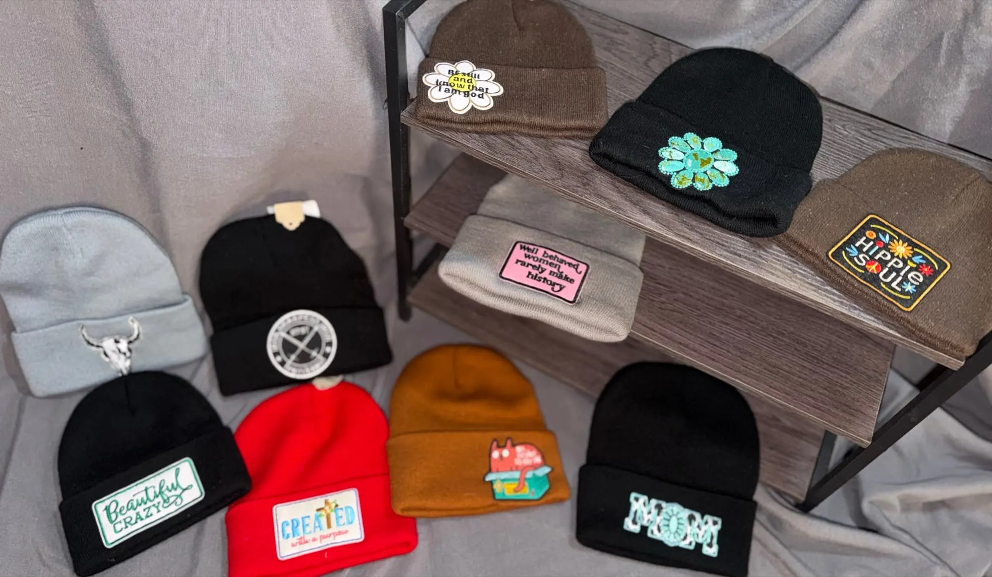 Ten patch beanies on and around a rack: Be Still and Know, a turquoise concho, Hippie Soul, a bull skull, a circle logo, Well Behaved Women, Beautiful Crazy, Created With a Purpose, a critter in a lunchbox and MOM