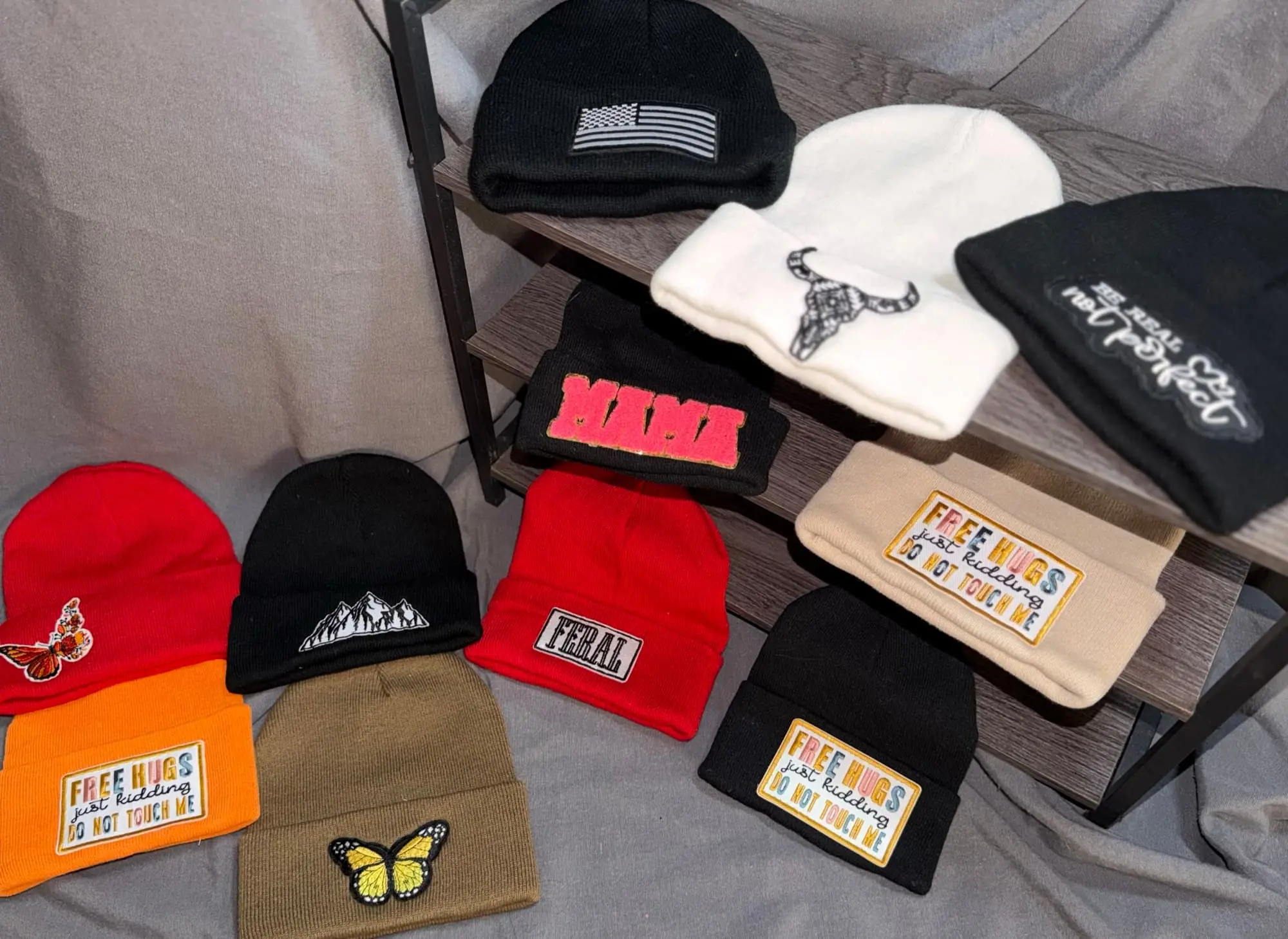 Eleven patch beanies: an American flag, a bull skull, Be Real Not Perfect, MAMA, three Free Hugs in tan, orange and black, a monarch butterfly, mountains, FERAL and a yellow butterfly