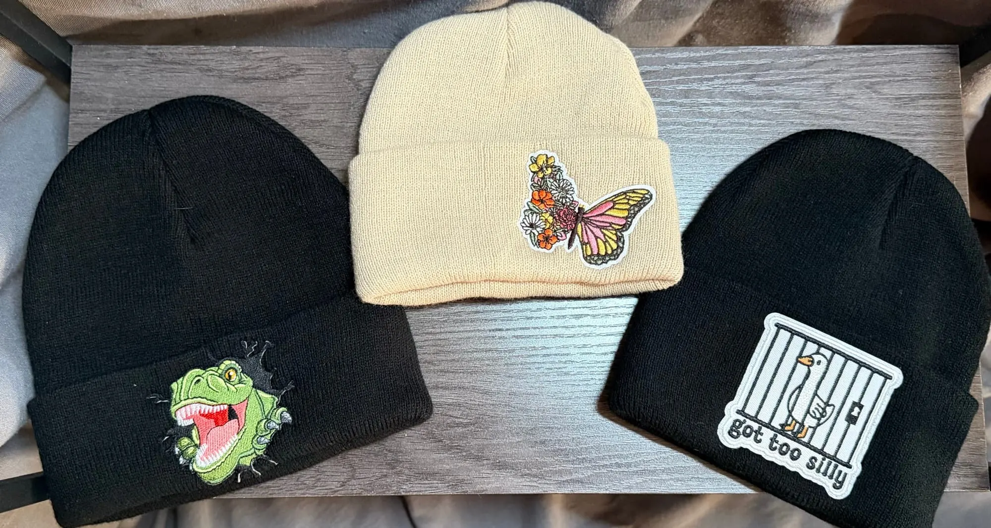 Three knit beanies laid flat: a T-rex bursting through black, a floral butterfly on cream and a got too silly goose on black