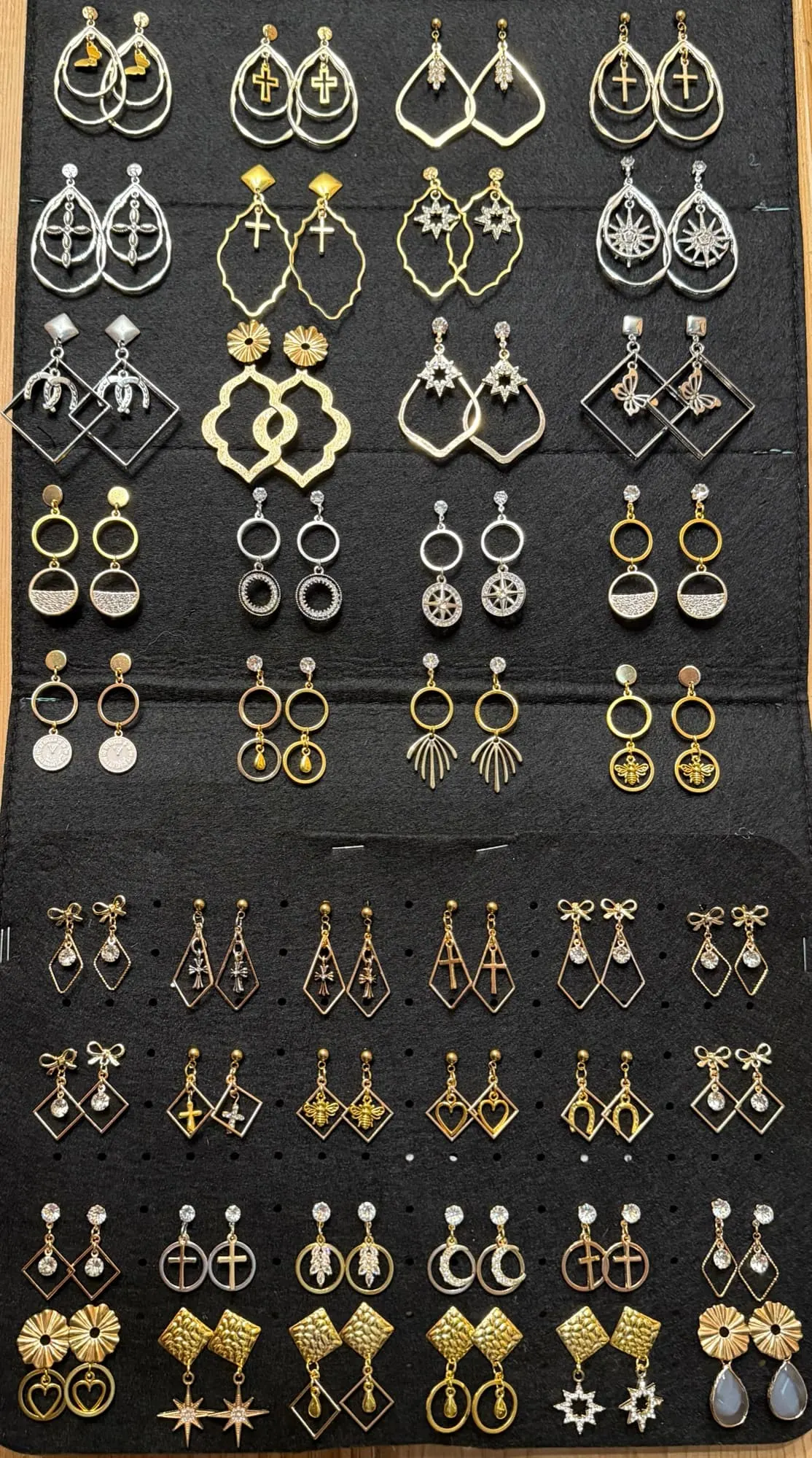 A black felt board filled with gold- and silver-tone earrings: teardrop hoops with crosses, starburst dangles, horseshoe and diamond drops, pavé circles and hammered studs