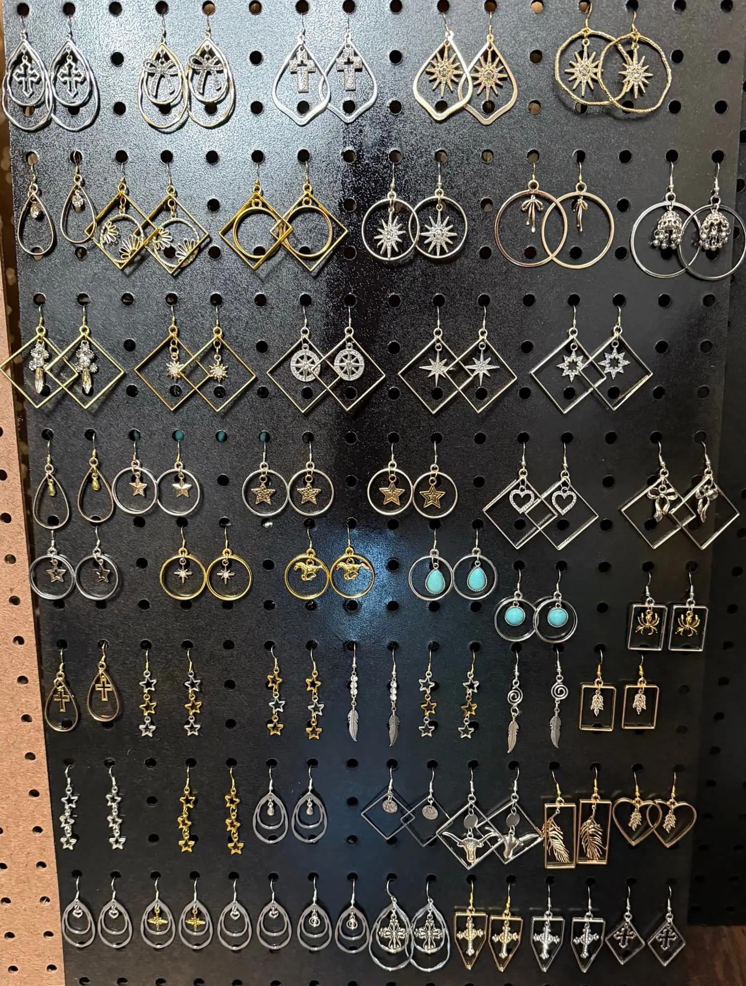 A black pegboard wall of silver- and gold-tone earrings: cross hoops, starbursts, stars, turquoise drops, feathers, longhorns and crosses