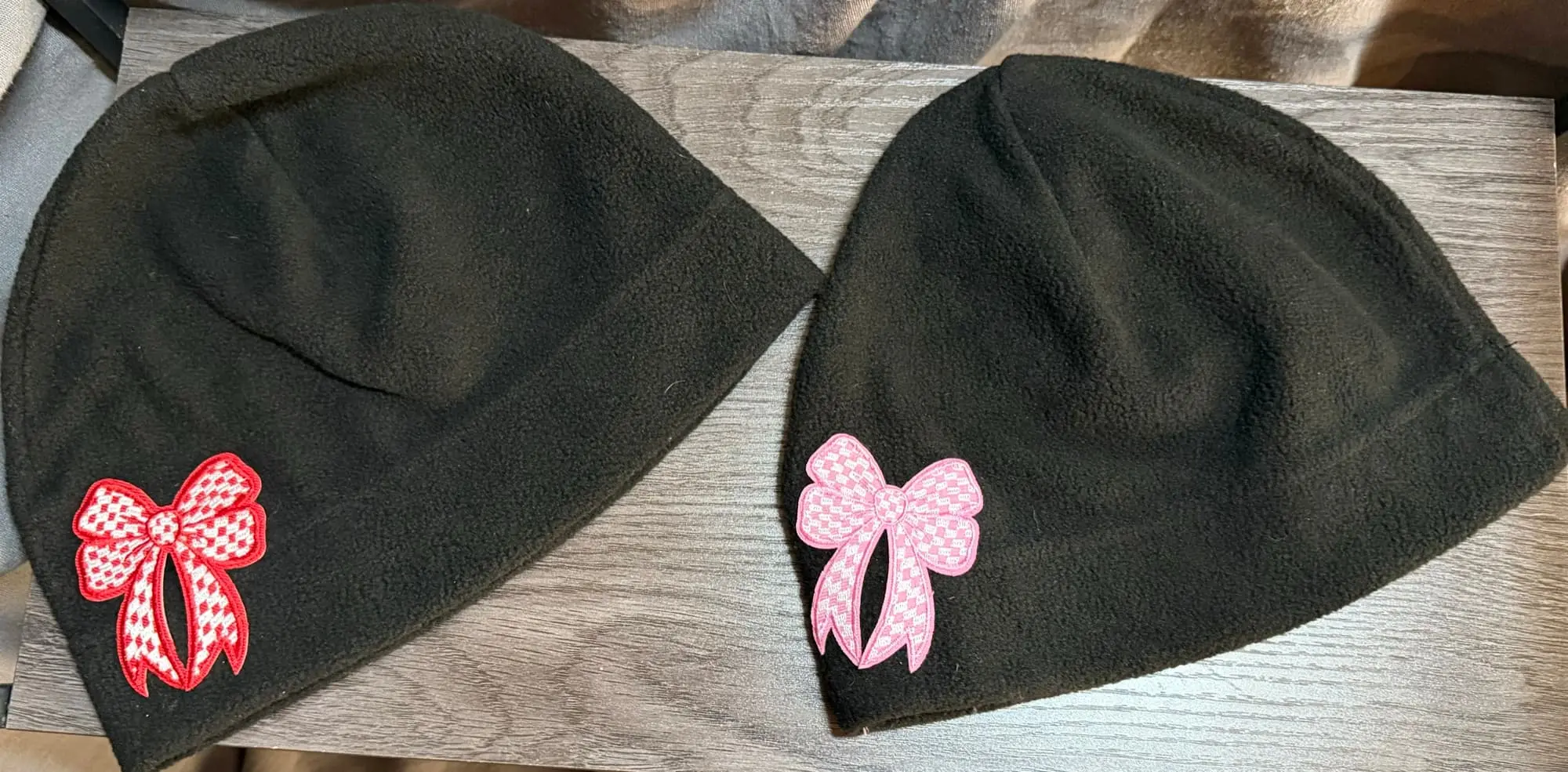 Two black fleece beanies laid flat, one with a red gingham bow and one with a pink gingham bow