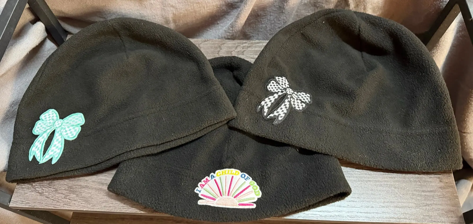 Three black fleece beanies: a teal gingham bow, I Am a Child of God with a sunrise, and a black-and-white gingham bow