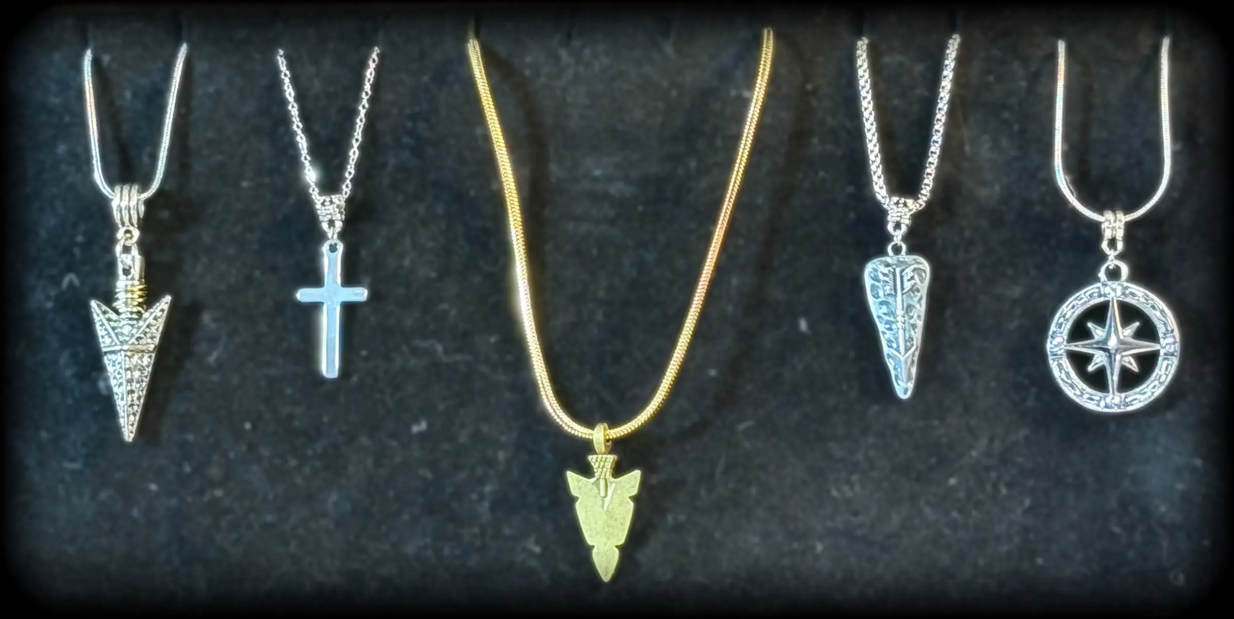 Five pendants: a silver arrowhead, a simple cross, a gold arrowhead, a tooled arrow charm and a compass rose
