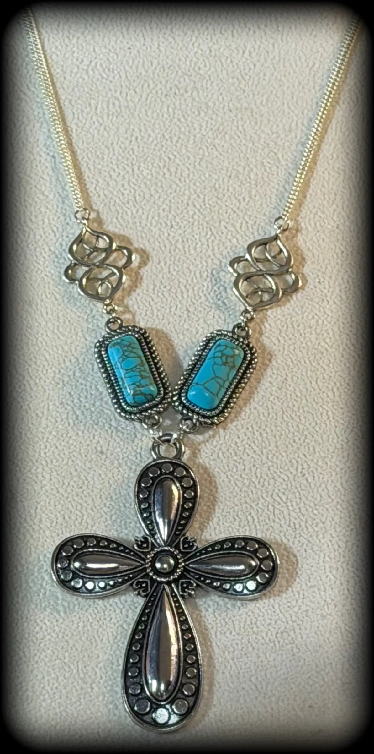 A necklace with turquoise bars and a black cross pendant on a cream display bust