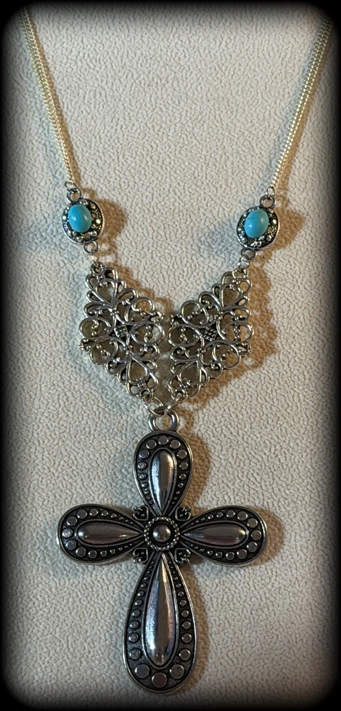 A necklace with turquoise accents, a filigree V and a black cross pendant on a cream display bust