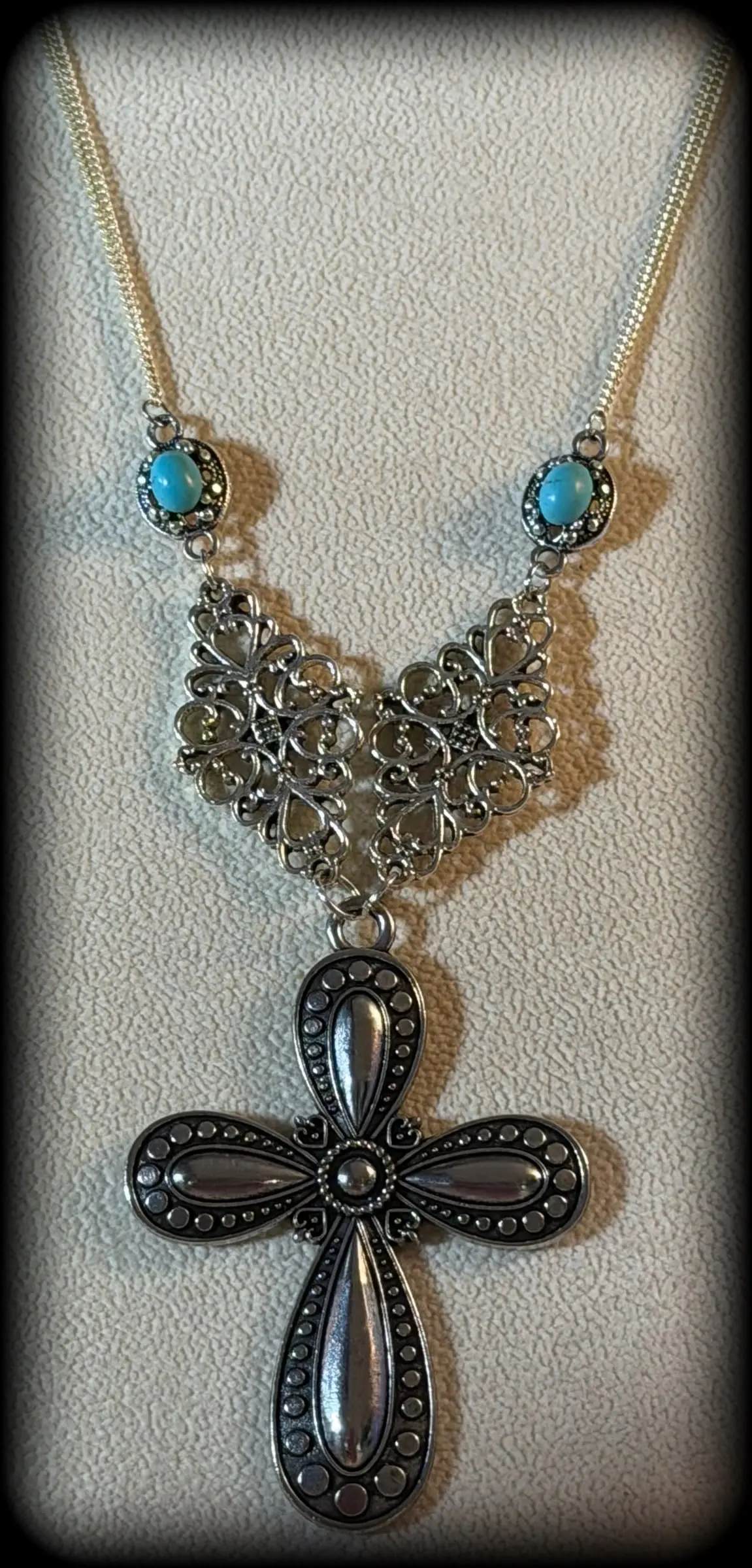 A necklace with turquoise accents, a filigree V and a black cross pendant on a cream display bust
