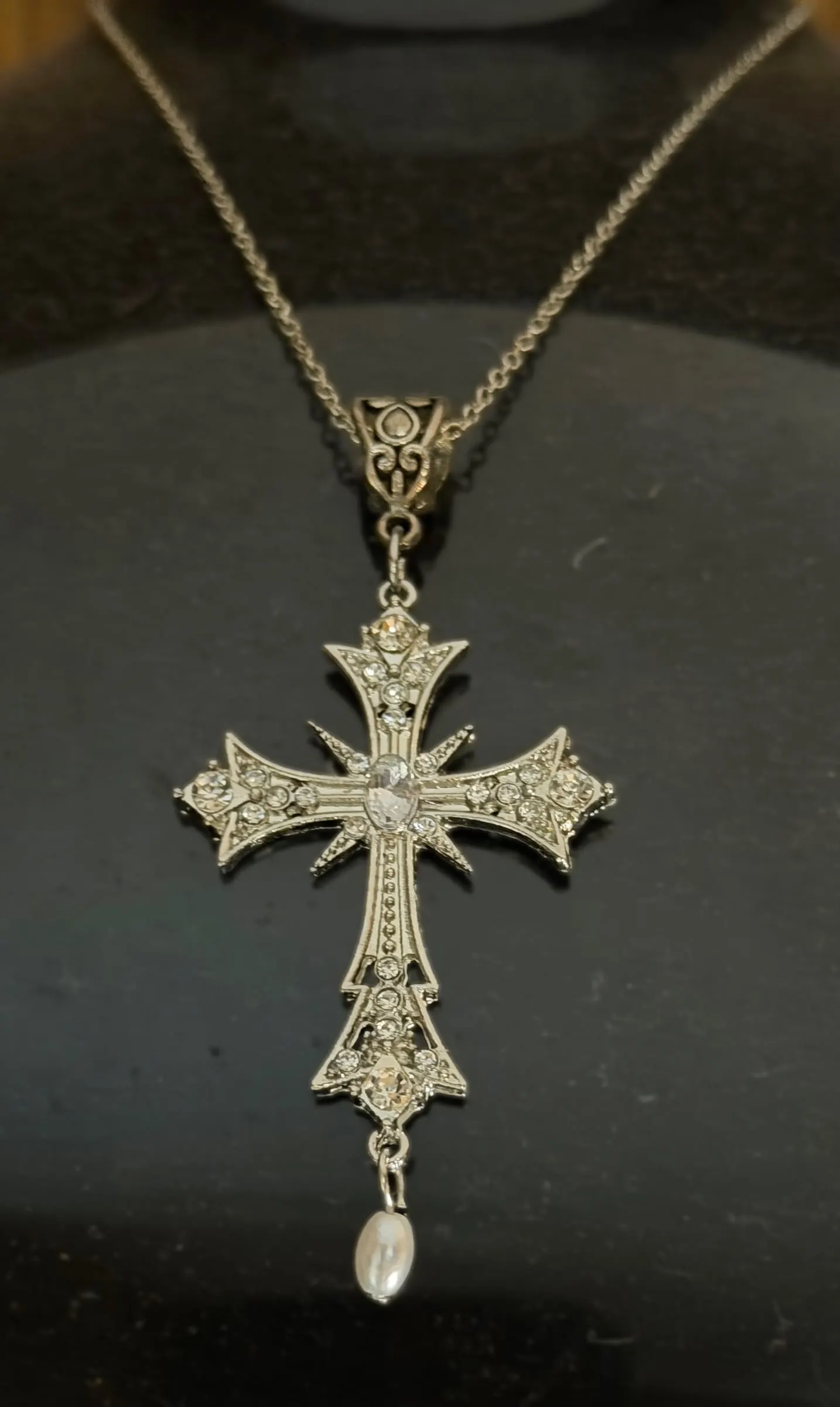An ornate silver filigree cross with a pearl drop on a black display