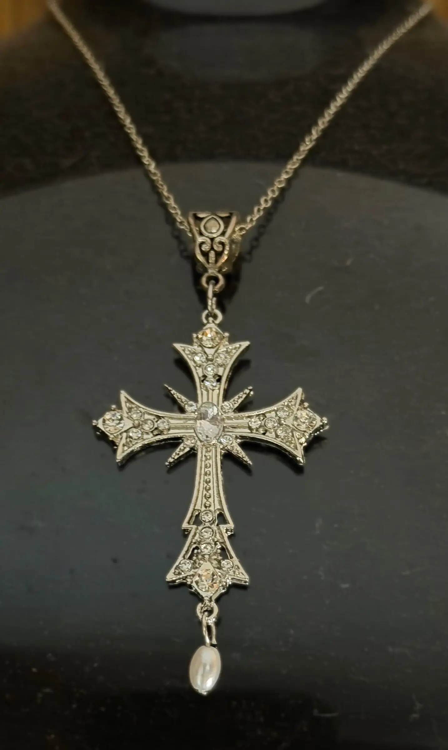 An ornate silver filigree cross with a pearl drop on a black display