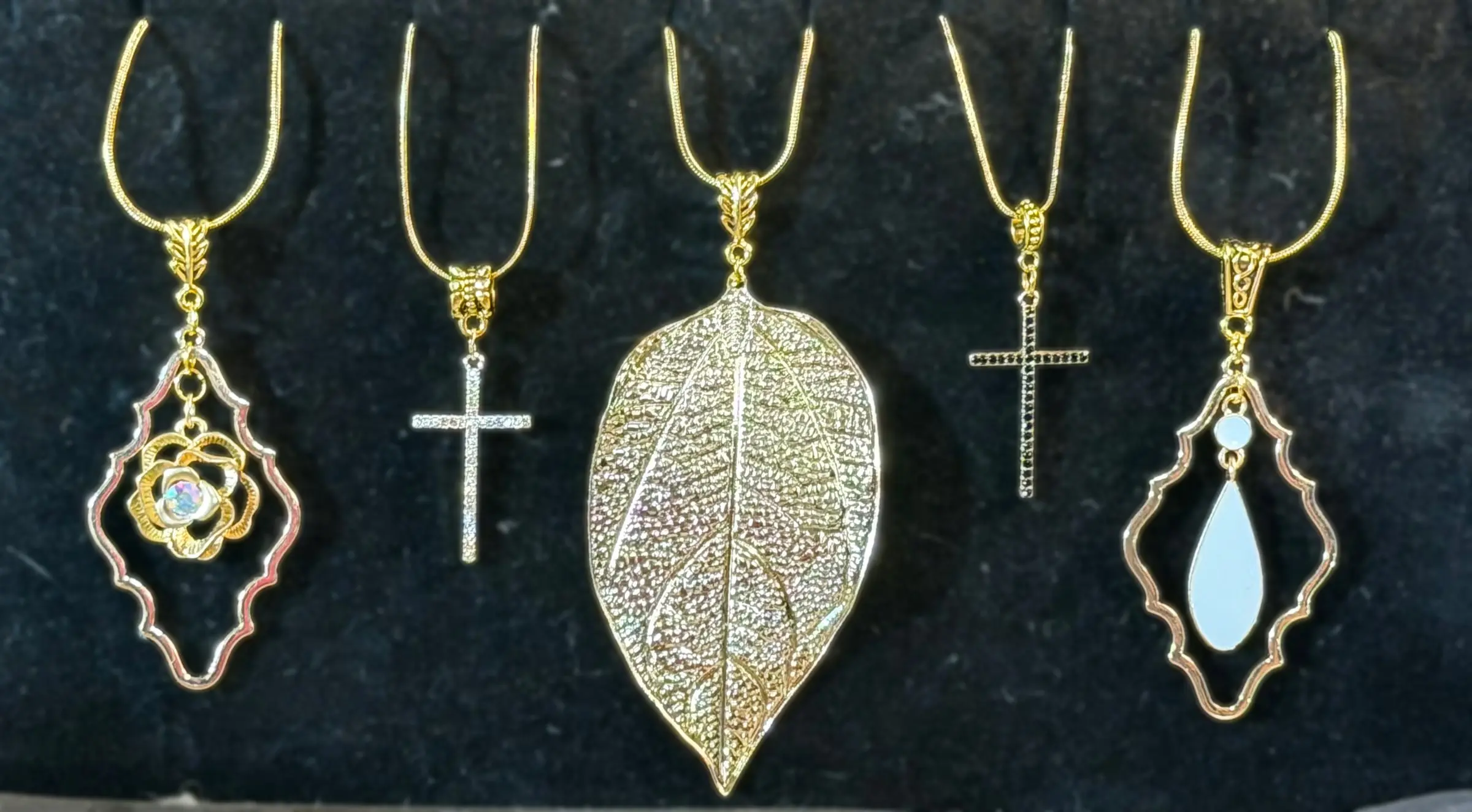 Five gold-tone pendants: a rose in a frame, a crystal bar cross, a gold leaf, a black crystal cross and an opal teardrop