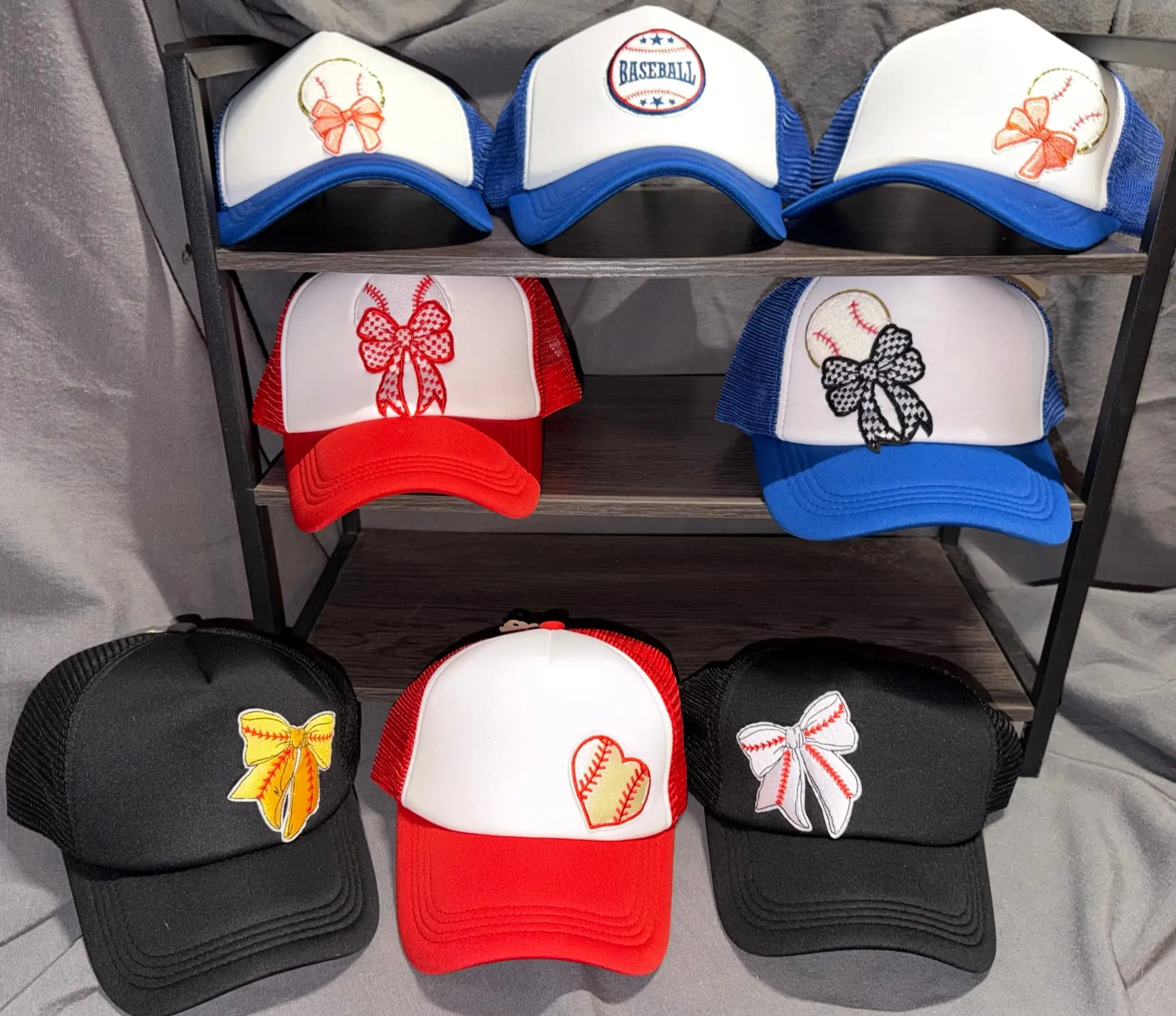 Eight baseball- and softball-themed trucker hats on a rack, with stitched bows, a Baseball badge and a baseball heart