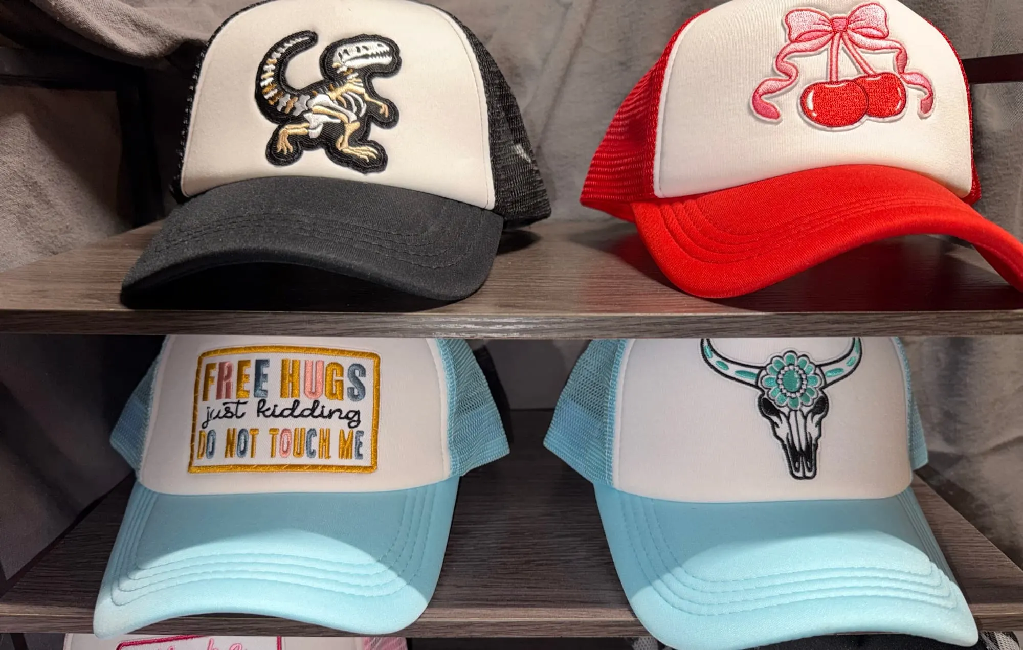 Four trucker hats on a wooden rack: a dinosaur skeleton on black, cherries with a pink bow on red, Free Hugs and a turquoise bull skull on light blue