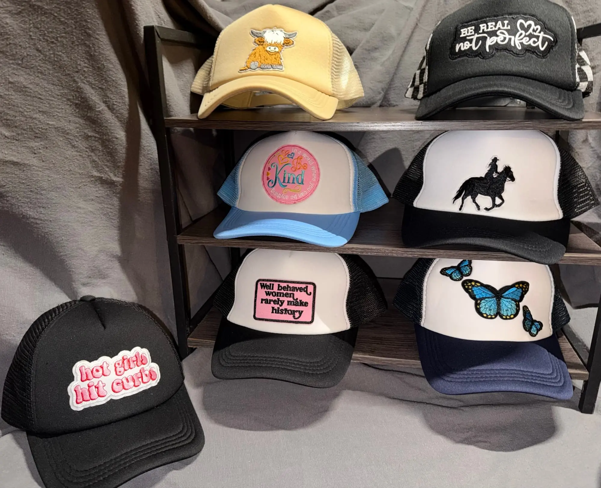 Seven trucker hats on a rack: a highland cow, Be Real Not Perfect, Kind, a horse and rider, Well Behaved Women Rarely Make History, blue butterflies and Hot Girls Hit Curbs