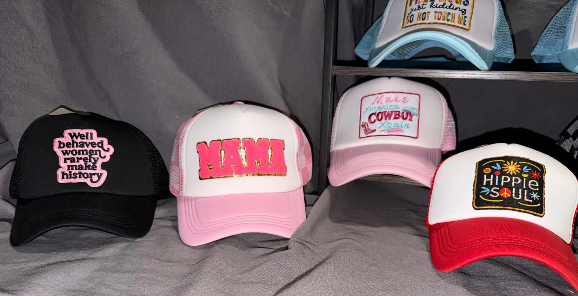 Four trucker hats: Well Behaved Women Rarely Make History on black, a pink chenille MAMA, Make America Cowboy Again in pink, and Hippie Soul in red