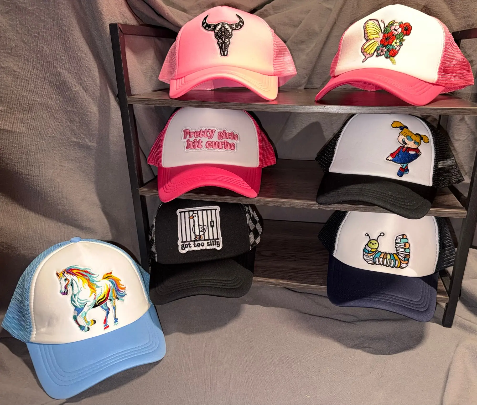Seven trucker hats on a rack: a pink bull skull, a butterfly bouquet, Pretty Girls Hit Curbs, a cartoon girl, a goose behind bars reading got too silly, a rainbow horse and a caterpillar