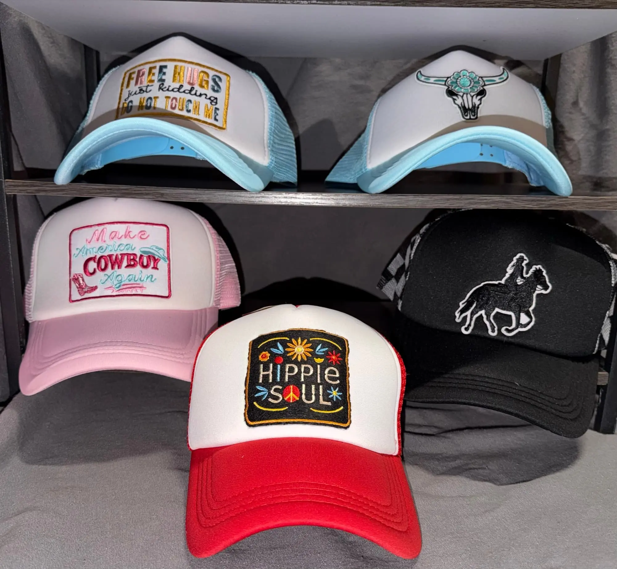 Five embroidered-patch trucker hats on a rack: Free Hugs, a turquoise bull skull, Make America Cowboy Again, Hippie Soul and a horse-and-rider silhouette