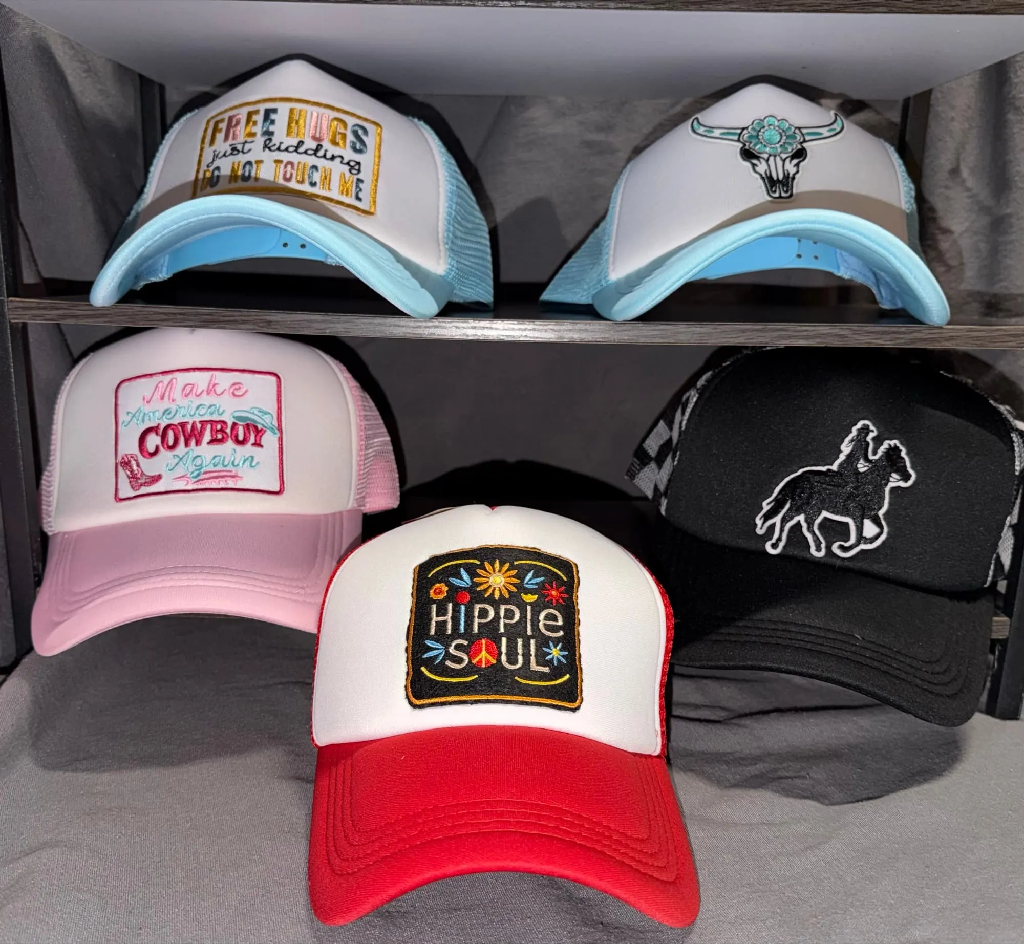 Five embroidered-patch trucker hats on a rack: Free Hugs, a turquoise bull skull, Make America Cowboy Again, Hippie Soul and a horse-and-rider silhouette